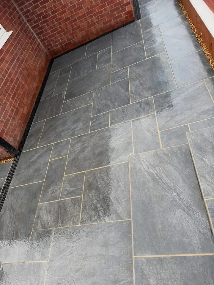 Completed grey stone-effect patio laid in a random pattern, with a black drainage channel running along the red brick wall of the house.