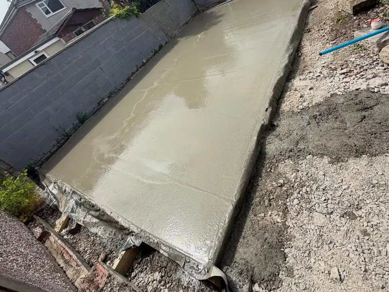 Freshly laid concrete base running along a garden boundary wall, with a compacted stone edge beside it.
