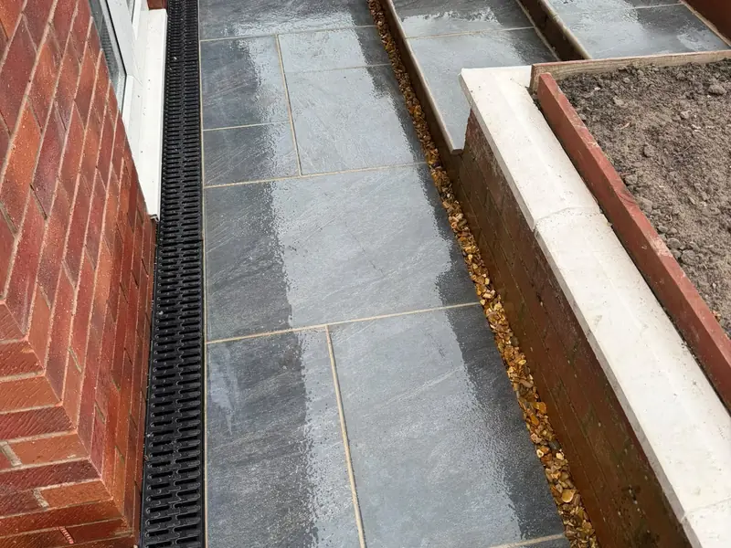 Grey paving beside a house wall with a channel drain running along its edge and steps up to a raised bed.