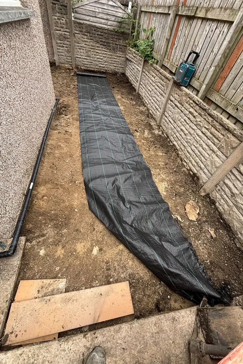 A narrow side garden dug out and levelled, with a membrane laid along the excavated ground.