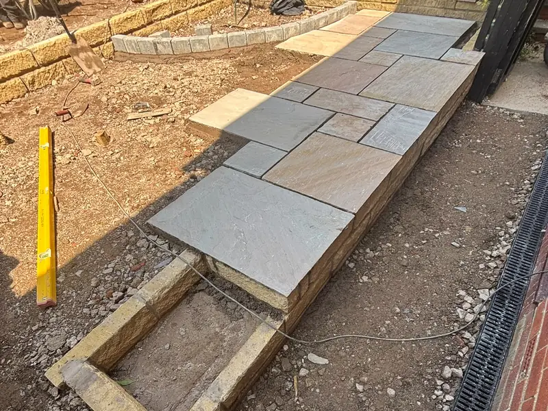 New paving being laid on a prepared base, with a channel drain along the house wall.