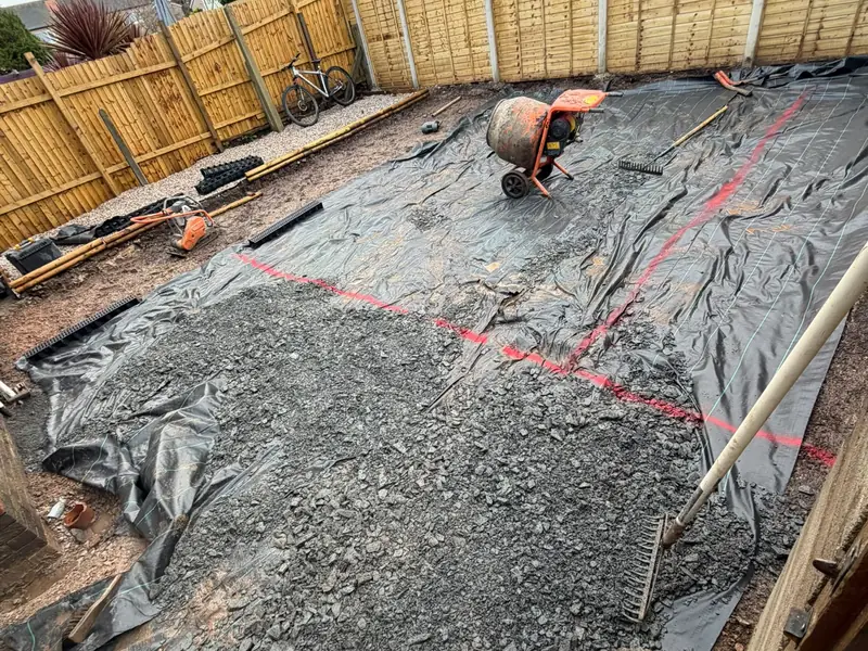 A large garden area prepared for new paving, with crushed stone sub-base spread over a membrane and a cement mixer on site.