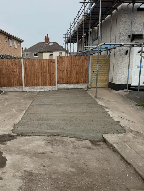 Timber fence panels and a side gate beside a house, with a freshly laid concrete area in front.