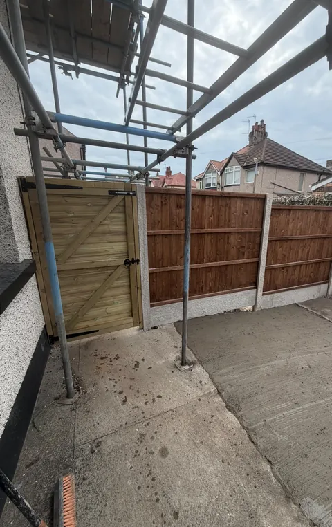 A ledged and braced timber side gate next to the fence, seen under scaffolding.