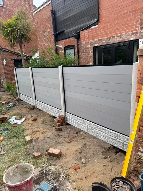 The first fence panels in place between concrete posts, with tools and bricks on the ground.
