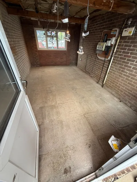 Inside the garage with bare brick walls, exposed ceiling joists, loose wiring and a concrete floor.