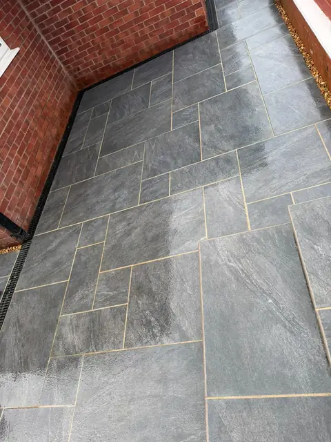 Grey paving beside a red brick house wall, with a channel drain along the edge.