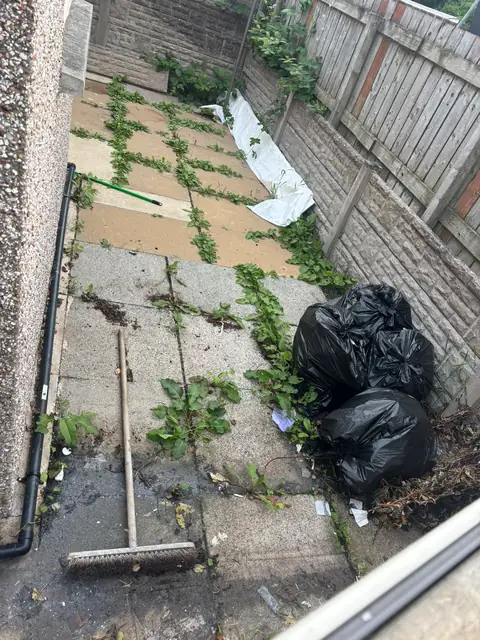 Old, cracked paving in a side garden, overgrown with weeds and cluttered with rubbish bags.