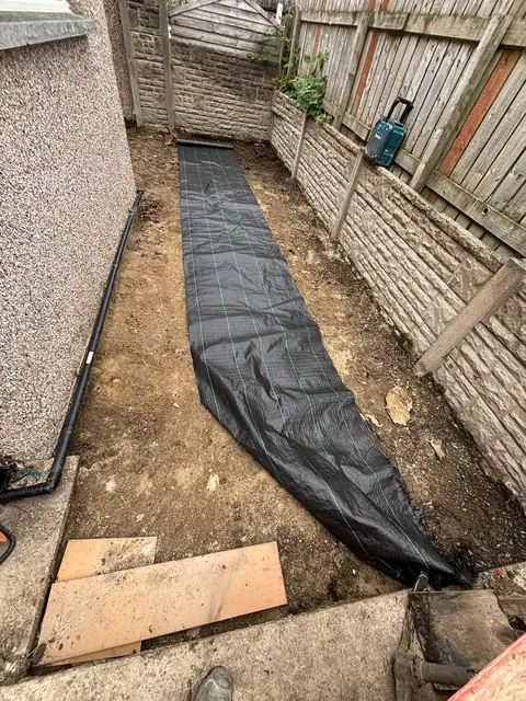 A narrow side garden dug out and levelled, with a membrane laid along the excavated ground.