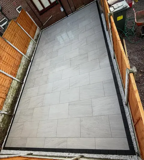 Light grey porcelain paving laid in a long, narrow garden between timber fences, with a dark border.