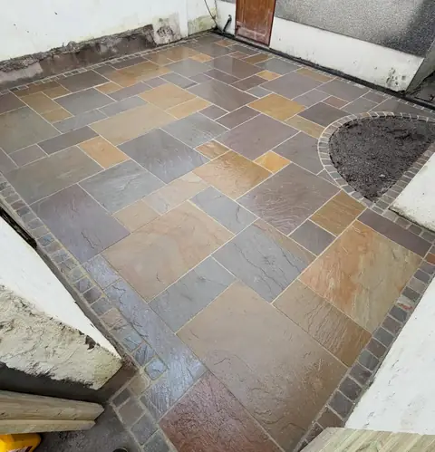 Indian stone patio in mixed buff, grey and rust tones, edged with a dark block border and a curved planting bed.