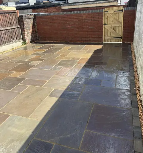 Completed patio in mixed-colour paving across a rear yard, with a red brick wall and timber gate.
