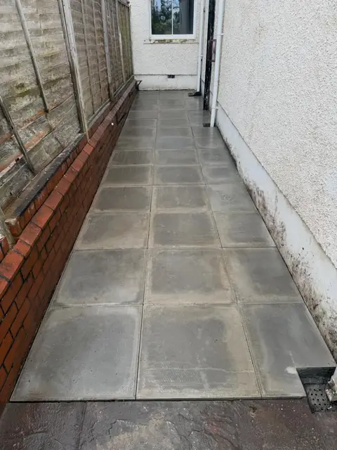 Grey paved side passage between a low brick wall and the side of a house.