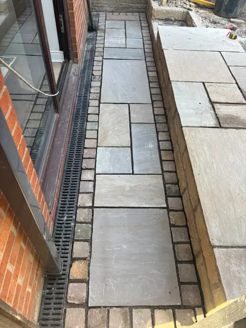 Narrow stone-paved path edged with setts, between a channel drain and a raised paved level.