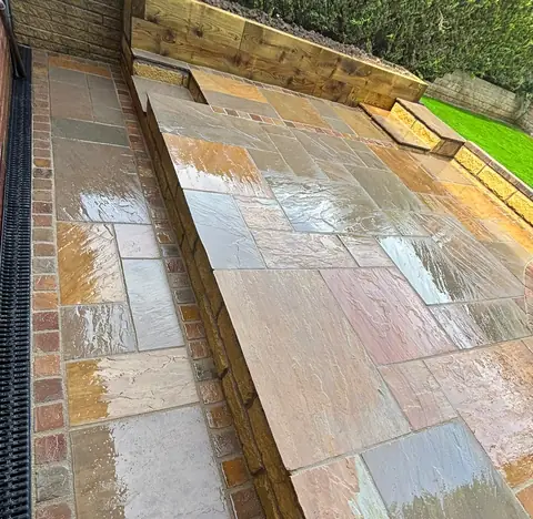 Finished paving with a drainage channel beside the house, a raised timber sleeper bed and artificial grass beyond