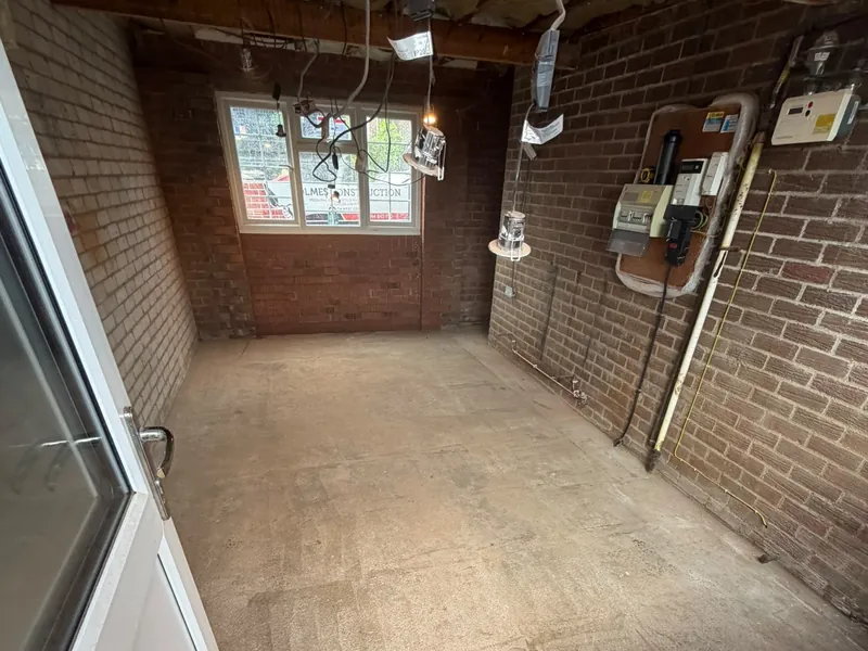 Inside a garage before the fit-out, with bare brick walls, exposed ceiling joists, loose wiring and a concrete floor.