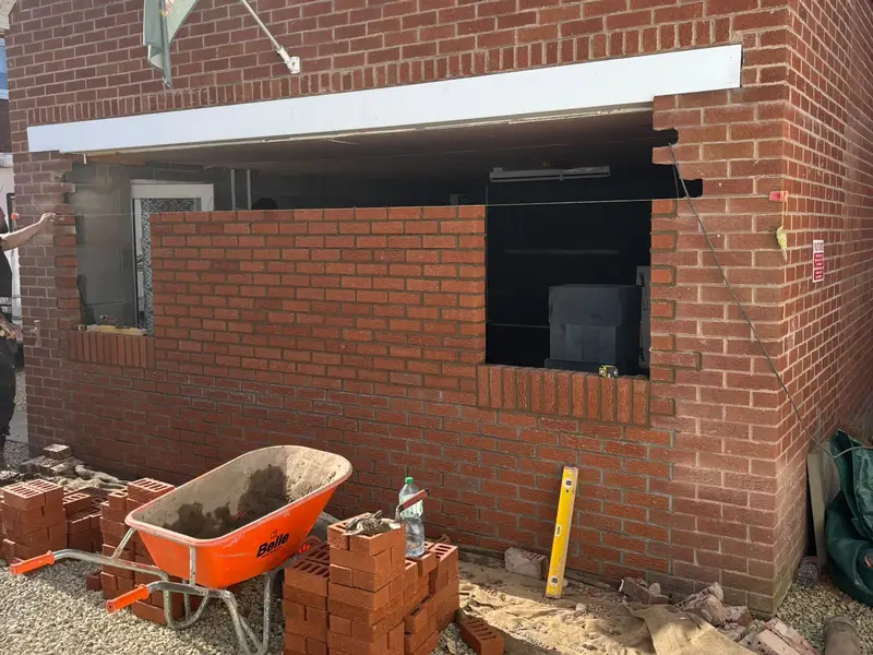Garage conversion underway, with the new brick front wall partly built and bricks and a wheelbarrow on the driveway.