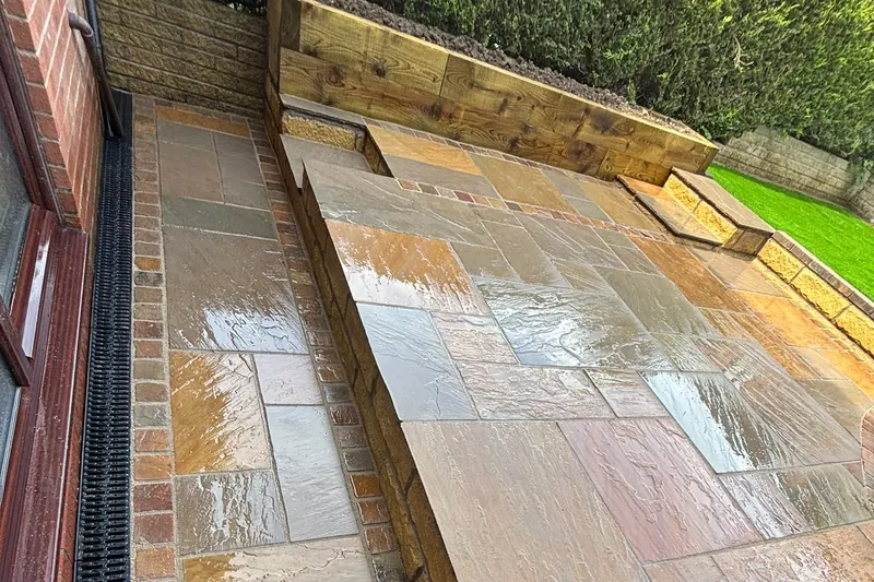Natural stone patio on two levels beside a house, with a channel drain, sett edging, a step and a timber sleeper raised bed.