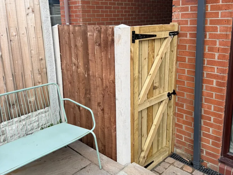 A timber side gate with black hinges beside a fence panel and concrete post, next to a brick house.