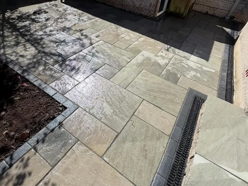 Rear garden with new natural stone paving, a channel drain and a planting border edged in dark blocks.