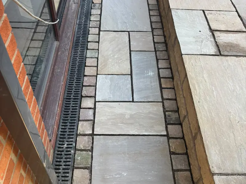 Narrow stone-paved path edged with setts, running between a drainage channel and a raised paved level.