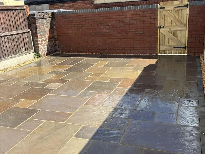 Completed patio in mixed-colour paving slabs, laid across a rear yard enclosed by a red brick wall with a timber gate.