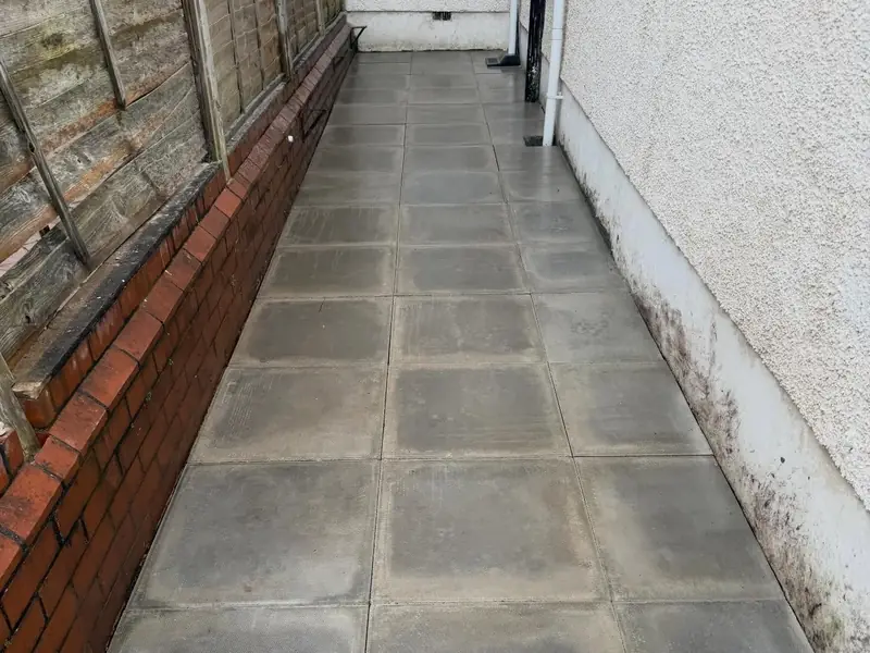 Grey paved side passage running between a low brick wall and the side of a house.