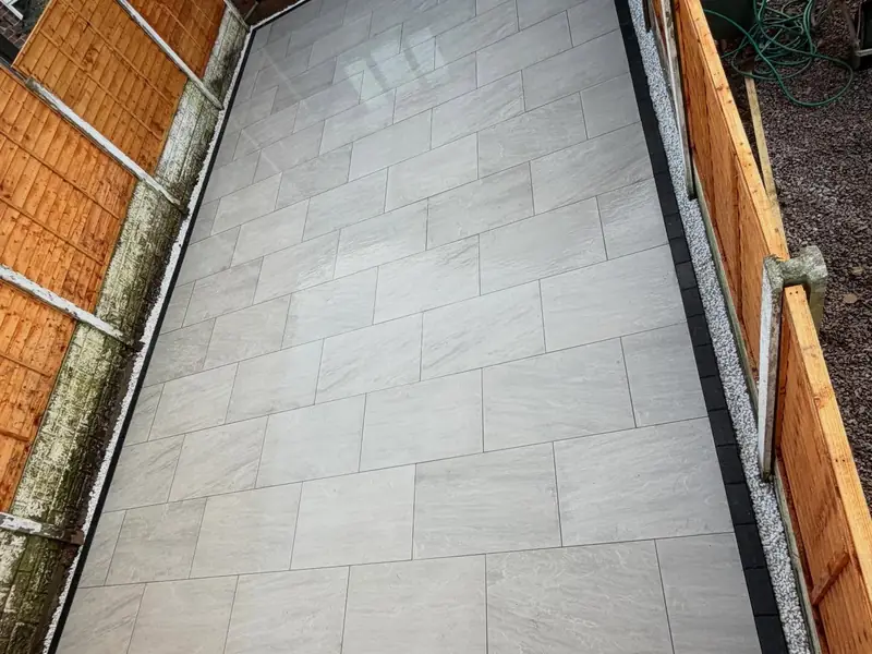Light grey porcelain paving laid in a long, narrow garden between timber fences, with a dark border along the edge.